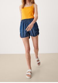 Canottiera gialla a coste, pantaloni shorts in denim blu con elastico in vita e sandali beige con cinturini regolabili. Design minimalista.