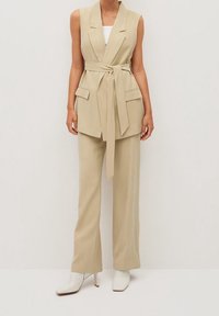 Beżowa kamizelka dopasowana z pasem, z dwiema kieszeniami z przodu. W połączeniu z dopasowanymi spodniami o szerokich nogawkach i białymi botkami na obcasie.