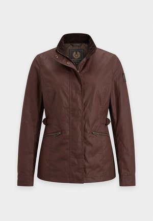 Femme portant une veste marron sur une chemise beige, un pantalon marron à jambes larges et des chaussures marron, debout avec les mains détendues le long du corps.
