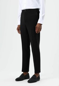 Zwarte, op maat gemaakte broek met een elastische tailleband, zijzakken en een recht gesneden pijp, gecombineerd met donkere loafers en een wit overhemd met knopen.