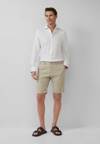 Weißes Leinen-Hemd mit Knöpfen, beiger Karottenschaden und braunen Sandalen. Mit hochgekrempelten Ärmeln und Taschen an den Shorts. Sauberer, moderner Schnitt.