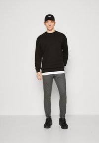 Felpa nera con colletto a girocollo, pantaloni grigi slim-fit e sneakers nere. Un berretto nero completa l'outfit, con un logo sulla parte anteriore.