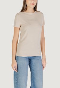 T-shirt in cotone beige con maniche corte. Presenta il logo "GUESS" in lettere embossate sul davanti. Abbinato a jeans in denim blu chiaro.
