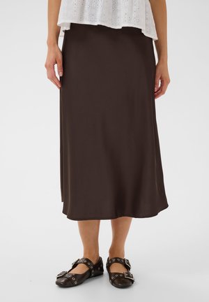 Žena nosiaca tmavohnedú midi sukňu, biely top s dierkami a čierne rovné topánky so sponami a kovovými detailmi, stojaca na jednoduchom pozadí.