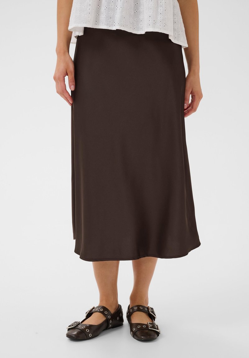Femme portant une jupe midi marron foncé, un top blanc à œillets et des chaussures plates noires avec boucles et œillets, debout sur un fond uni.