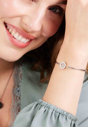 Zilveren bangle armband met een ronde hanger met een uitgesneden "B", gedragen om een pols. Zachte groene blouse met plooien in de mouwen.
