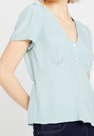 Vrouw draagt een lichtblauw, kort mouwens blouse met knopen en een V-hals, gecombineerd met donkerblauwe spijkerbroek, staande tegen een effen achtergrond.