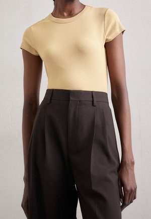 Osoba nosząca beżową prążkowaną bluzkę z krótkim rękawem, wpuszczoną w spodnie z wysokim stanem o szerokich nogawkach w ciemnym brązie na jednolitym tle.