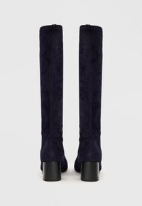 Bottes en daim bleu marine à hauteur de genoux avec un talon épais noir et sans matériel visible, présentant une texture lisse et un design ajusté.