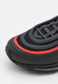 Black athletic shoe with red accents, featuring a textured mesh upper, curved design, and a visible lace-up system. Reinforced toe cap.