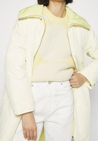 Chaqueta de crema acolchada con un cuello verde claro, combinada con un suéter suave de patrón amarillo y vaqueros blancos, mostrando una textura suave.