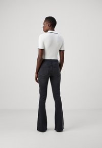 Top corto bianco a coste con bordo nero, abbinato a jeans a zampa scuri, visto da dietro su uno sfondo grigio chiaro.