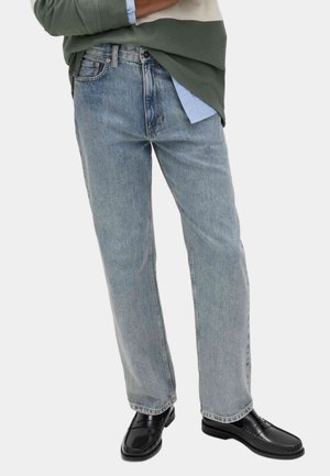 Persoon die lichtblauwe, losvallende jeans draagt, zwarte leren loafers en gelaagde groene en blauwe tops, staand met gekruiste armen.