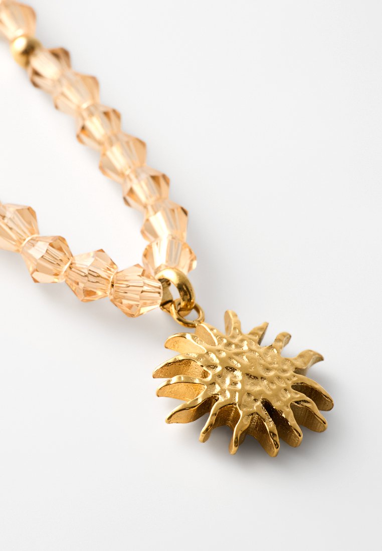 Goldener, sonnenförmiger Anhänger an einer Perlenkette mit hellen Pfirsich-Kristallperlen, der eine strukturierte Oberfläche und ein poliertes Finish aufweist.