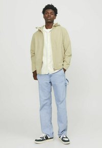 Lys beige zip-up jakke med hætte, lagd over en stribet skjorte med knapper, parret med lyseblå jeans og sorte sneakers.