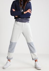 Pantaloni felpati a patchwork grigio chiaro con dettagli blu, orli a costina, realizzati in tessuto morbido. Indossati con una giacca bomber blu notte e sneakers bianche.