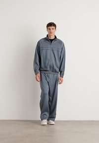Grå treningsdress laget av glatt stoff, med en jakke med glidelås og ståkrage samt bukser med elastisk midje. Bæres med hvite joggesko.