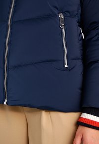 Granatowa kurtka puchowa z pikowanym wzorem, z metalowym suwakiem i rękawami ściągaczami w czerwono-białe paski. Ciepły i casualowy styl.