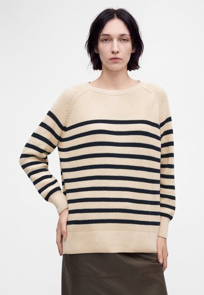 Femme aux cheveux courts et foncés portant un pull beige à rayures horizontales noires et une jupe foncée, debout devant un fond clair uni.