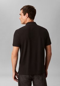 Man in zwart, kortärmlig poloshirt en donkere spijkerbroek staat met zijn gezicht van ons af tegen een effen grijze achtergrond.