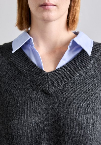 Pulover gri tricotat cu guler în V, suprapus peste o cămașă cu guler de culoare albastru deschis. Țesătura pare moale, având o textură ondulată aproape de decolteu.