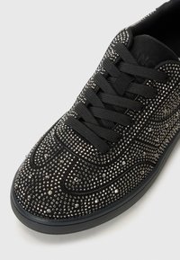 Mustad musta tennis jalanõud, millel on läikivad rhinestone-kivid, paksude paeltega ja tekstuuriga pinnaga, heleda halli tausta ees.