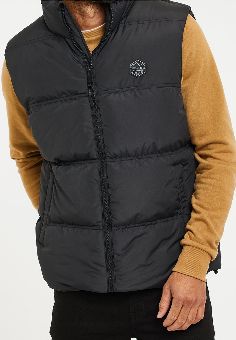 Homme portant une veste matelassée noire zippée avec un logo sur la poitrine, par-dessus une chemise marron à manches longues, debout, les mains détendues le long du corps.