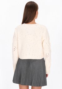 Lichtromige gebreide trui met een cropped ontwerp, voorzien van spikkels en openwerk accenten, gecombineerd met een geplooide grijze rok.