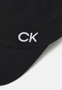 Juodas minkštos medžiagos kepurė su išlenktu skydeliu ir išsiuvinėtu baltu "CK" logotipu priekyje, skirtu ženklinimui.