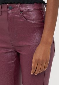 Jeans bordeaux brilhantes com um corte slim. Apresentam um bolso frontal, fecho com botão e um leve brilho. Textura suave com um brilho subtil.