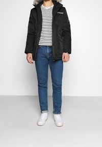 Parka negra con capucha forrada de piel, camiseta a rayas, vaqueros azules y zapatillas blancas; cuenta con cierres de cremallera y botón.