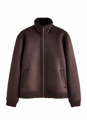 Personne portant une veste marron avec une main placée dans une poche latérale, montrant les poignets et le bas côtelés.