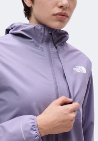 Personne portant une veste North Face violet clair, tenant la fermeture éclair près de la poitrine, avec la capuche de la veste visible.