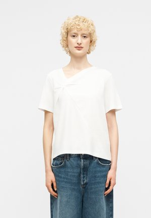 Νεαρό άτομο με κοντά σγουρά ξανθά μαλλιά που φορά λευκό κοντομάνικο τοπ και μπλε τζιν παντελόνι με φαρδιές μπροστινές παντελόνες μπροστά σε απλό φόντο.