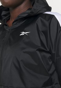 Gros plan d'une personne portant une veste noire à capuche zippée avec un logo blanc sur la poitrine et une bande blanche sur l'épaule.