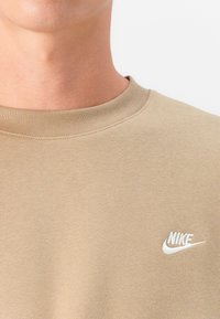 Beige sweatshirt van zacht materiaal, met een ronde halslijn en een wit geborduurd Nike-logo op de linkerbovenborst.