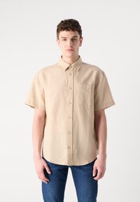 Camicia beige a maniche corte con bottoni sul davanti, colletto e taschino sul lato sinistro del petto. Realizzata in tessuto leggero con una texture liscia.