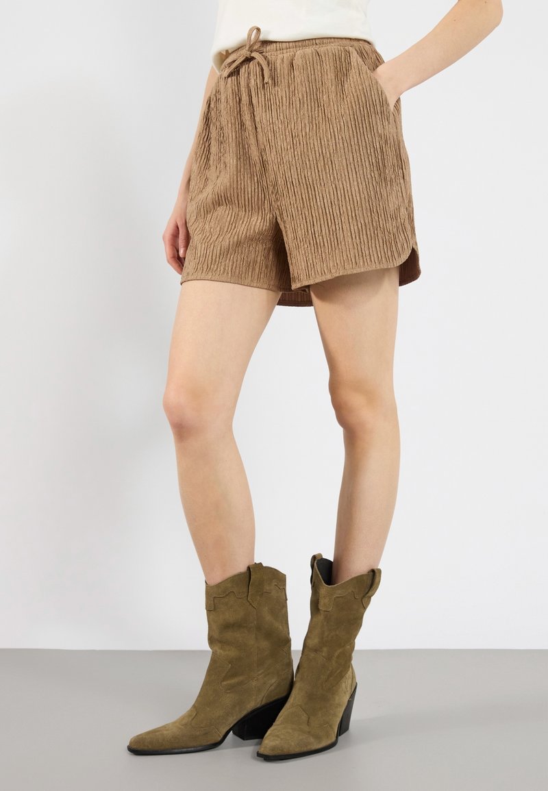 Personne portant un short marron texturé avec un cordon de serrage et des bottes de cow-boy en daim vert, debout sur un fond clair uni.