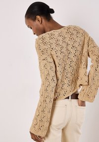 Beige gestrickter Pullover mit offenem Spitzenmuster, langen Ärmeln und lockerer Passform, kombiniert mit hellen Hosent.