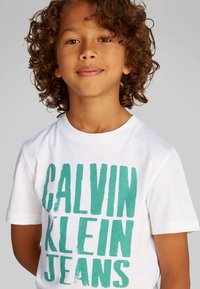 Wit katoenen T-shirt met groene blokletters met de tekst "CALVIN KLEIN JEANS." Korte mouwen en een ronde hals. Gladde textuur.