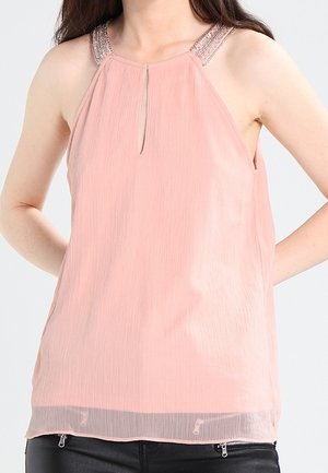 Vrouw draagt een mouwloze lichtroze blouse met kralenversiering bij de halterhals en een keyhole-detail, gecombineerd met een zwarte broek met ritsen.