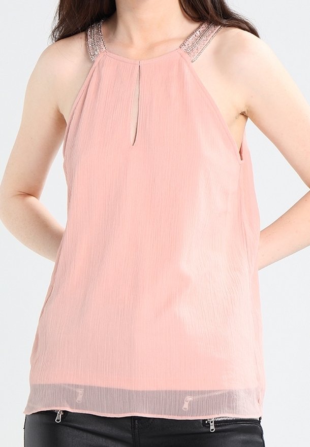 Femme portant un chemisier sans manches rose clair avec un col halter orné de perles et un détail œillet, associé à un pantalon noir zippé.