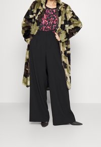 Faux-Fur-Mantel im Camouflage-Muster, über einem floralem Top; hoch taillierte schwarze weite Hosen; schwarze spitz zulaufende Schuhe.