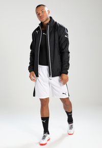Sort letvægtsjakke med hvid detaljer, båret over en sort top, parret med hvide shorts og sorte sokker. Funktionen Puma-branding gennemgående.