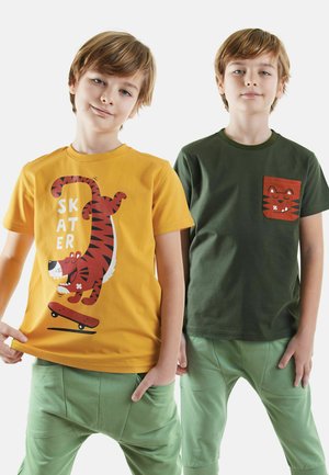 To drenge bærer legesyge T-shirts: den ene i orange med et tiger grafik og tekst, den anden i olivengrøn med et katte-lomme design. Begge har grønne bukser.