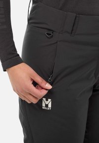 Schwarze Softshell-Hose mit einer Reißverschlusstasche, die ein kleines Logo am Oberschenkel und eine strukturierte Stoffoberfläche aufweist. Eine Hand greift den Reißverschluss.