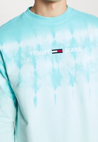 Jasnoniebieska bluza tie-dye z logo "TOMMY JEANS" w białym kolorze oraz małym wzorem flagi w kolorach czerwonym, białym i niebieskim. Długie rękawy, tkanina z mieszanki bawełny.