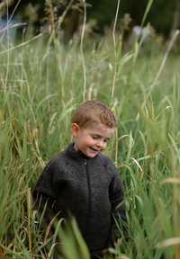 Barn i en mörkgrå fleecejacka med hög krage, omgiven av högt grönt gräs. Mjuk textur, dragkedja framtill, ledig passform.