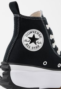 Sneaker alta nera in canvas con lacci bianchi e suola in gomma; presenta un logo circolare con una stella nera e il testo "Converse All Star".