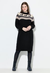 Schwarzes gestricktes Kleid mit einem gemusterten beigen Ausschnitt, der geometrische Designs aufweist. Das Model trägt dunkle Strumpfhosen und schwarze Schnürstiefel.